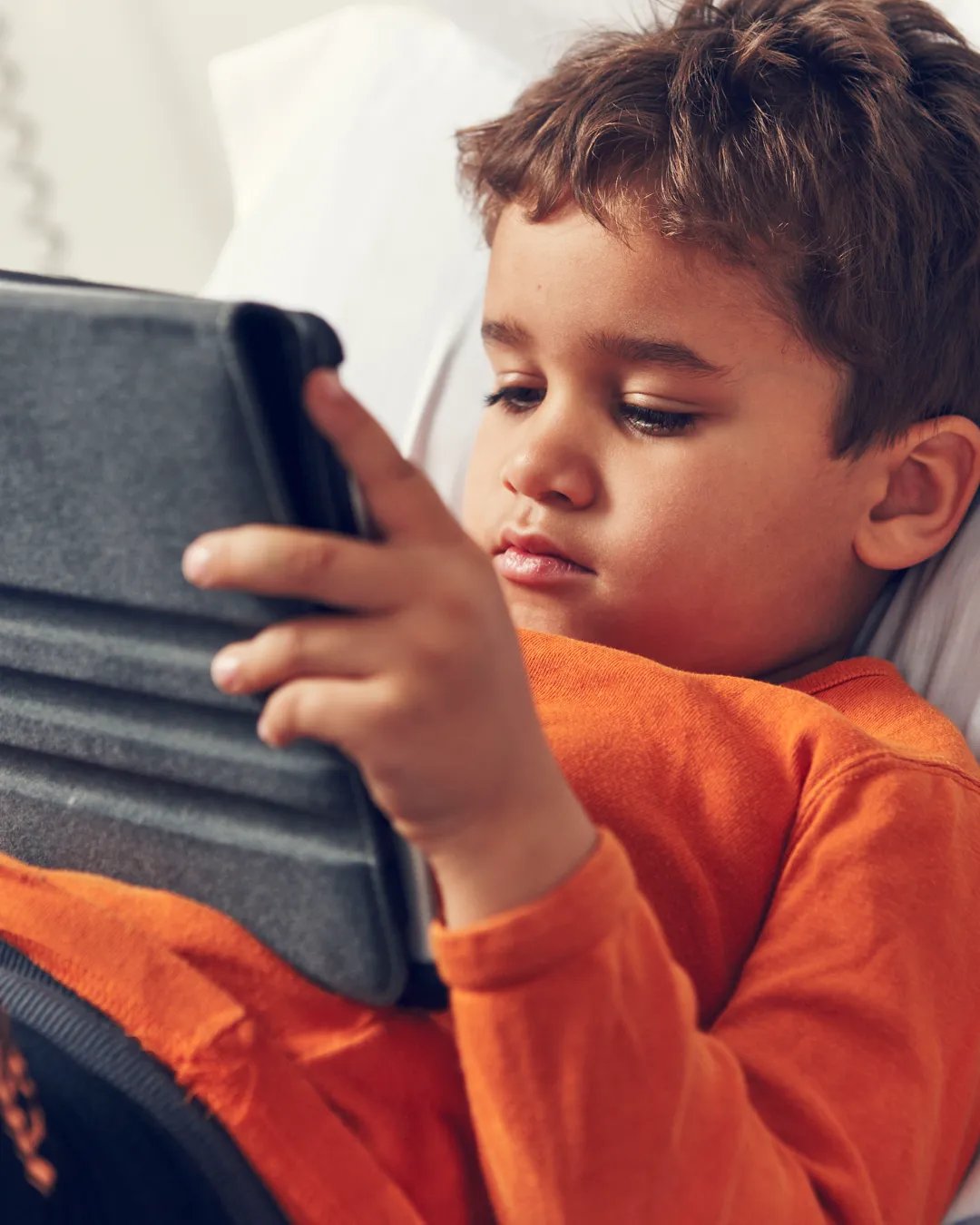 A young boy laying in bed looking at a tablet.